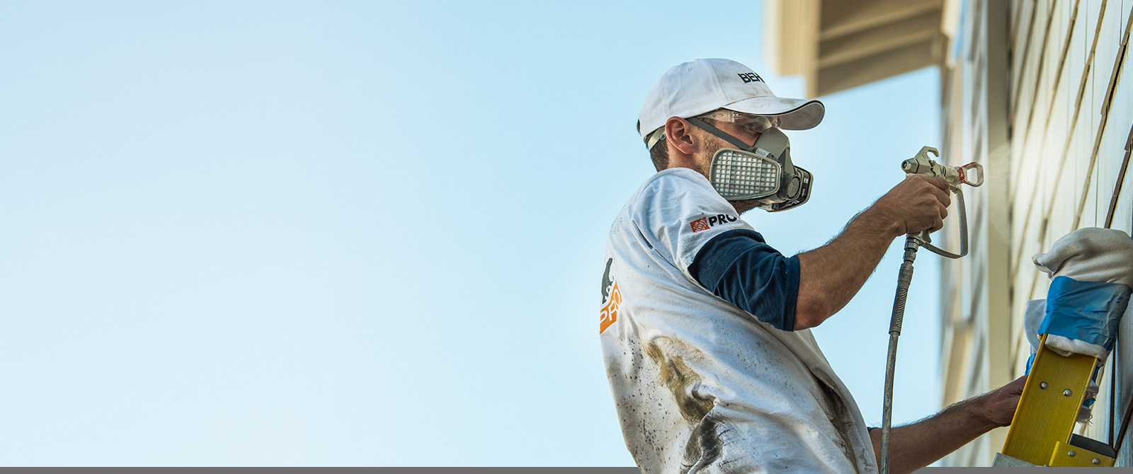 Image of a Pro Contractor wearing a hat and shirt with Behr logo spray painting an exterior wall of a house on a ladder.