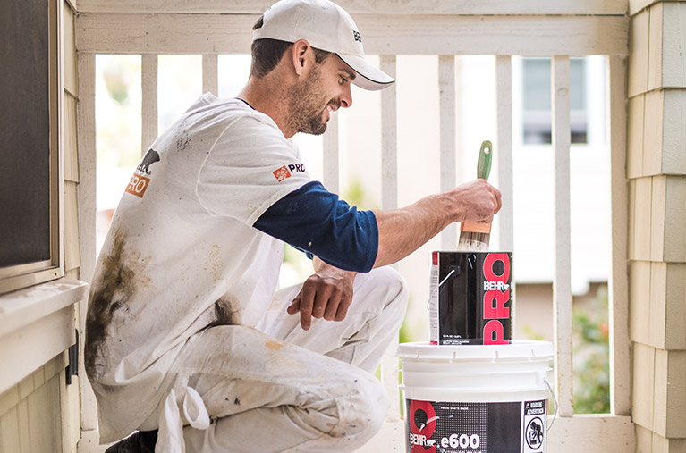 A mobile view of a Pro Contractor wearing a cap and shirt with Behr logo dipping a paint brush on a 1 gallon of BEHR PRO e600 paint can on a porch that he is going to paint.