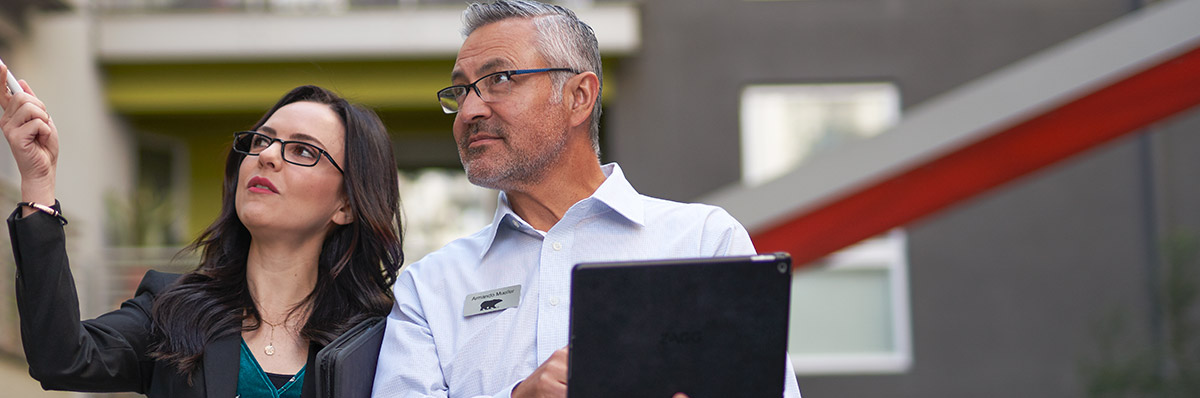 Large image a property manager and a BEHR PRO Rep looking at what the property manager is pointing at on the outside of an apartment complex. The BEHR PRO rep is taking notes on an IPAD
