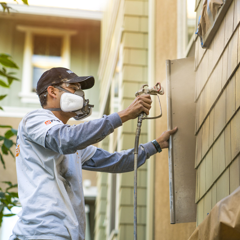 Mobile view of a pro painter spray painting the exterior wall of a house.