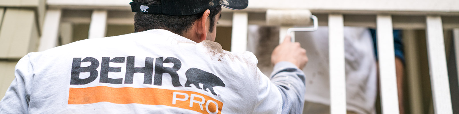 A close-up image of a back of a painting contractor who is painting with a roller with the logo of BEHR PRO imprinted on the shirt..