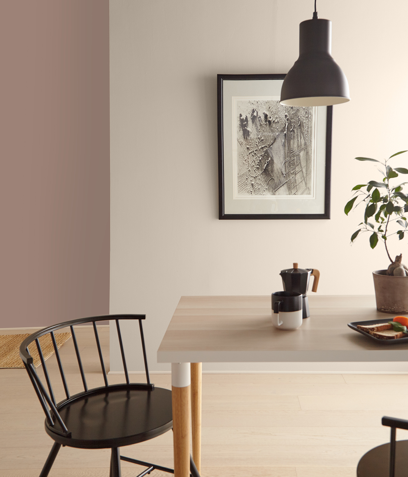 A casual dining setting showing a corner of a table and chair. On the table is coffee, plate of food and potted plant. A light hangs down above the table.