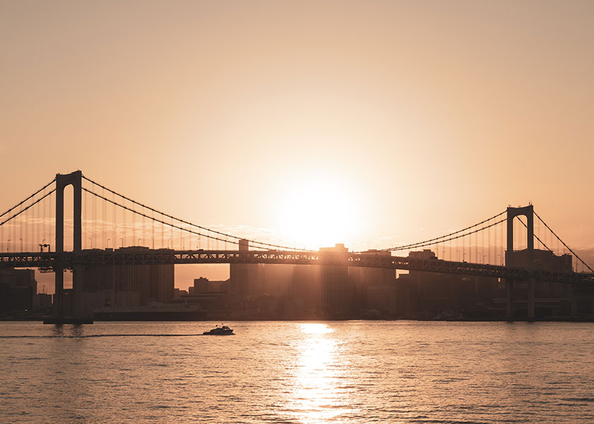 A pulled back view of a bridge that sits over a body of water. The sun is setting and glowing in the sky.