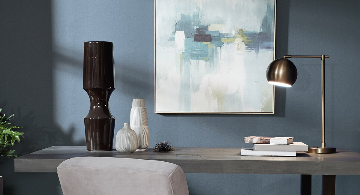 A calming office with a graycolored wood desk sitting against a wall painted in blue. There is a abstract painting hanging on the wall.