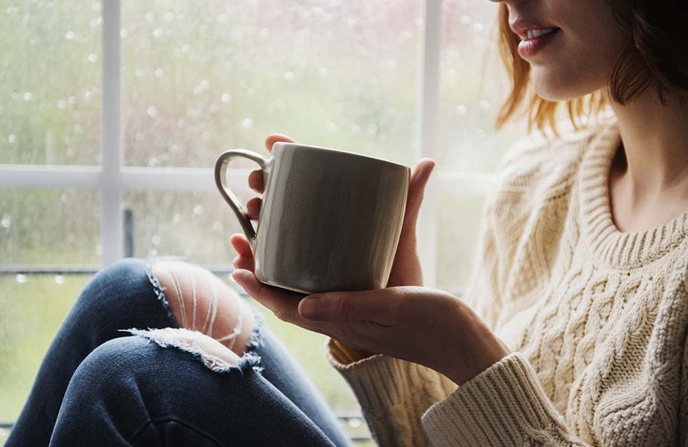 A woman sitting cozily by a window comforted by a warm cup of coffee. Outside is raining.